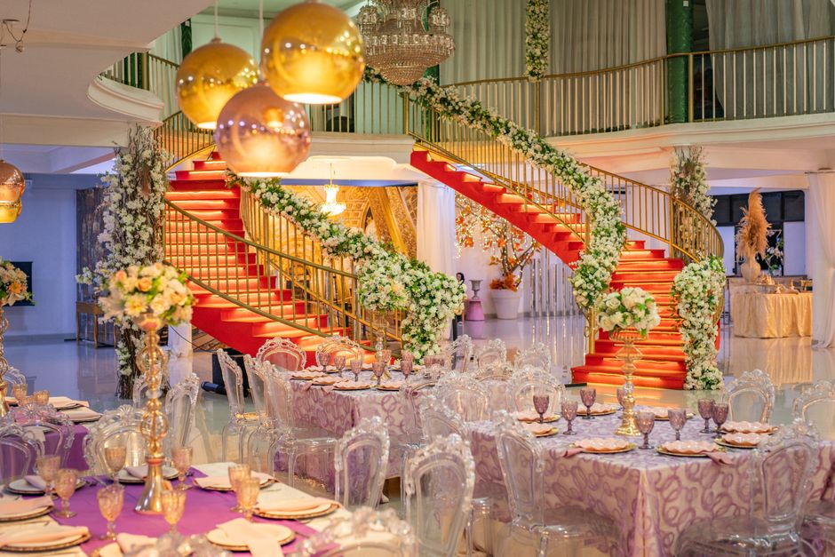 A luxurious floral arrangement featuring white roses, peonies, and greenery, elegantly draped along the sweeping staircase with red carpeting in an opulent event venue. The staircase is adorned with a