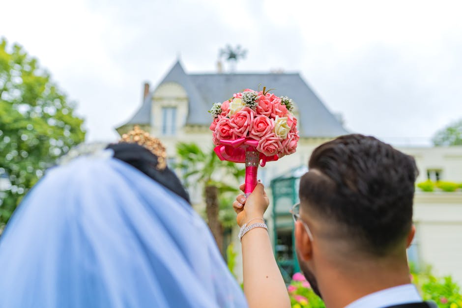 A close-up of a person holding a round floral bouquet arrangement featuring pink and white roses, with small white filler flowers, wrapped with a pink ribbon. The bouquet is held in front of a bride d