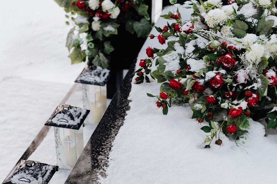 A memorial arrangement of red rose bouquets with green leaves and some white flowers, partially covered in snow, laid on a black marble ledge at a cemetery. Small white candles in glass holders, with
