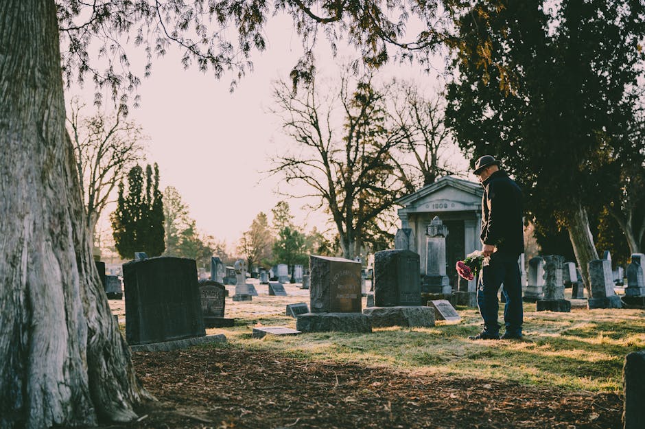 A man dressed in dark clothing, including a hat, stands in a cemetery holding a floral arrangement of pink and purple flowers. Surrounding him are various gravestones and memorials, some engraved with