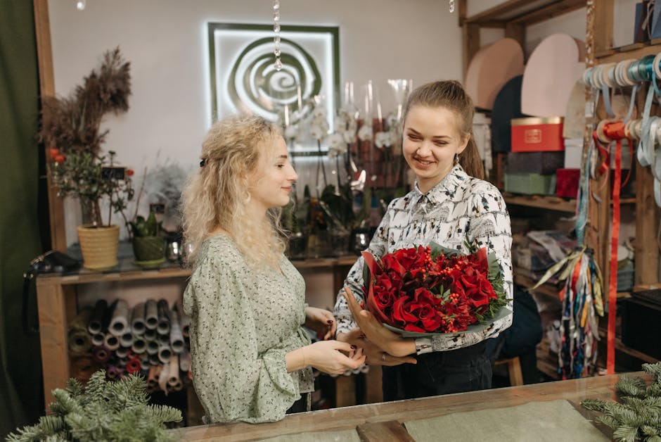 A young woman with light skin and long, dark hair tied back in a ponytail, dressed in a patterned blouse, is holding a vibrant floral bouquet featuring red roses, lilies, and greenery, wrapped in gree