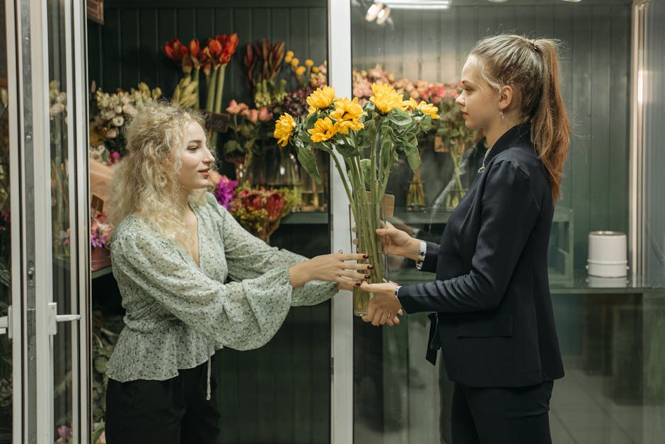 A floral delivery scene inside a flower shop features two women exchanging a bouquet of vibrant yellow and orange sunflowers with green foliage. The woman on the left, with curly blonde hair, is weari