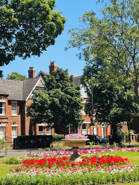 The image shows a bright, sunny day in a well-maintained city park with lush green trees and a clear blue sky. In the foreground, there is an ornate stone planter filled with pink and purple flowers,